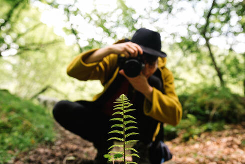 Talentierte Fotografin in trendigem Outfit, die eine Pflanze auf einem Feld mit einer Fotokamera fotografiert - ADSF17361