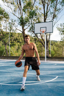 Konzentrierter männlicher Sportler mit nacktem Oberkörper, der allein auf einem Sportplatz im Sommer Basketball spielt - ADSF17387