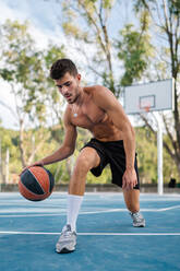 Concentrated male athlete with naked torso playing basketball alone on sports ground in summer - ADSF17388