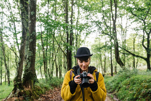 Weibliche Fotografin in modischer Kleidung steht auf einem Weg im grünen Wald und macht ein Foto - ADSF17392