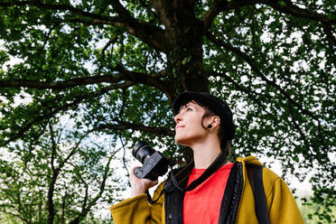 Weibliche Fotografin in modischer Kleidung steht auf einem Weg im grünen Wald und macht ein Foto - ADSF17395