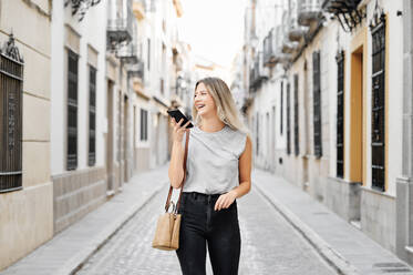 Happy young female in stylish outfit messaging on mobile phone while strolling on old narrow street in city looking away - ADSF17416