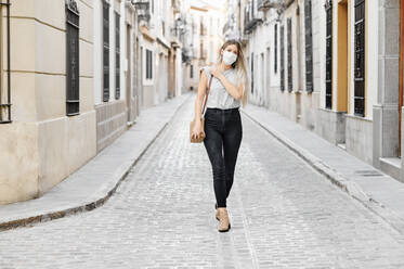 Young female in stylish outfit wearing a face medical mask walking on the street looking away - ADSF17417