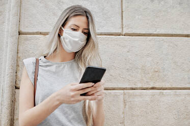 Young female in stylish outfit wearing a face medical mask messaging on mobile phone while standing against old stone concrete building city - ADSF17418