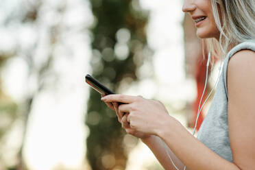 Side view of happy young female in casual outfit listening to music with earphones and browsing smartphone while spending summer day on street - ADSF17420