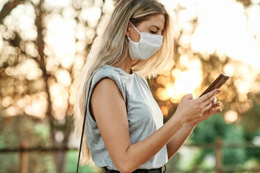 From below cheerful young female in trendy outfit and protective mask for coronavirus prevention browsing on smartphone while standing in green park in sunny day - ADSF17421
