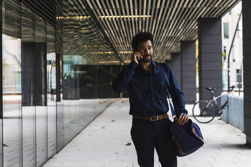 Nachdenklicher Unternehmer mit Tasche, der in der Stadt mit seinem Smartphone telefoniert - PNAF00077