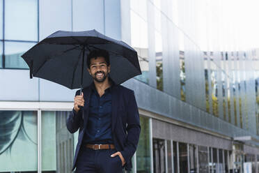 Lächelnder Unternehmer mit Hand in der Tasche, der einen Regenschirm gegen ein Gebäude in der Stadt hält - PNAF00081