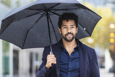 Lächelnder Unternehmer mit Regenschirm in der Stadt während der Regenzeit - PNAF00108