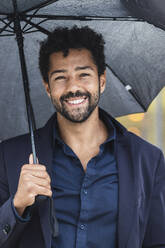 Lächelnder Unternehmer mit Regenschirm in der Stadt - PNAF00109