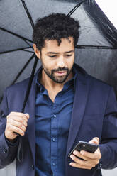 Unternehmerin mit Regenschirm und Smartphone in der Stadt - PNAF00110
