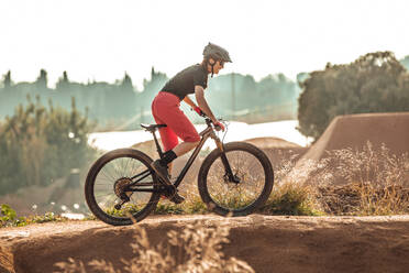 Seitenansicht einer Sportlerin in schwarzem Helm und roter Sportkleidung mit Brille, die auf einer Trainingsstrecke Mountainbike fährt - ADSF17425