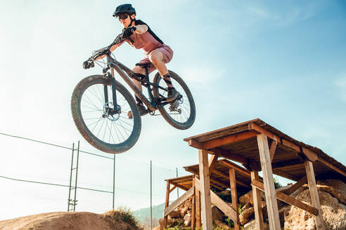 Sportlerin in schwarzem Helm und Sportkleidung mit Brille fährt Mountainbike und springt von einer Brücke im Trainingsgelände - ADSF17427