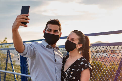 Lächelnder Freund und Freundin mit Maske, die auf einer Brücke stehen und ein Foto mit der Selfie-Kamera ihres Handys machen, während sie das Wochenende zusammen genießen - ADSF17457