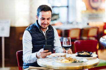 Lächelnder junger Mann, der im Restaurant sitzt und mit seinem Smartphone SMS schreibt - CJMF00352