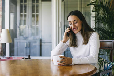 Lächelnde Frau mit Kaffeetasse, die mit ihrem Handy telefoniert, während sie zu Hause sitzt - ABZF03429