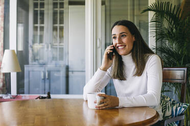 Junge Frau mit Kaffeetasse, die zu Hause sitzt und mit ihrem Handy telefoniert - ABZF03430