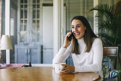 Young woman drinking coffee while talking on mobile phone at home stock photo