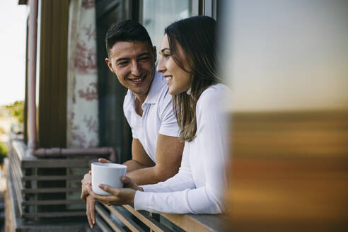Lächelnder Freund schaut Freundin an, die mit einer Kaffeetasse auf dem Balkon steht - ABZF03451