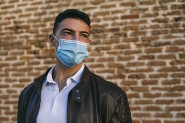 Young man wearing face mask standing against brick wall - ABZF03467