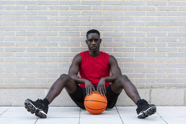 Selbstbewusster afroamerikanischer Basketballspieler in Sportkleidung, der auf einem Spielplatz mit einem Ball auf einem Steinrand sitzt und in die Kamera schaut - ADSF17469
