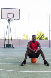 Ruhiger afroamerikanischer Sportler sitzt auf einem Basketball auf einem Spielplatz und schaut weg - ADSF17470