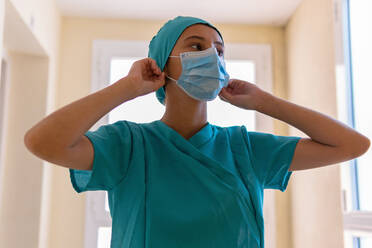 Ernste Krankenschwester in Uniform, die vor der Arbeit eine medizinische Maske anlegt, während sie in einer Klinik während einer Coronavirus-Epidemie steht und wegschaut - ADSF17476