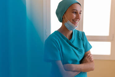 Positive young medical worker in uniform with mask standing near window and smiling happily while having break during work in hospital - ADSF17477