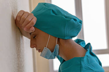 Side view of tired desperate female medic in blue uniform with mask on chin leaning on wall of hospital corridor after working hard during coronavirus pandemic - ADSF17482