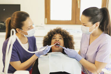 Weibliche Assistentin hält Absaugvorrichtung, während der Zahnarzt die Zähne einer afroamerikanischen Frau in der Klinik bohrt - ADSF17490