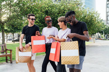 Fröhliche junge Studenten in trendigen Outfits und mit Sonnenbrillen, die ihre Einkäufe in Einkaufstaschen tragen und fröhlich plaudern, während sie eine Straße überqueren - ADSF17497