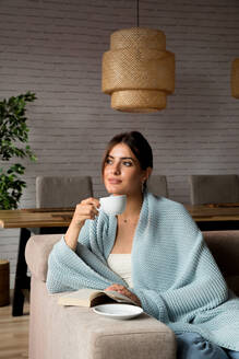 Nachdenkliche junge Frau, eingewickelt in eine warme Decke, sitzt auf der Couch und genießt einen heißen Tee, während sie im gemütlichen Wohnzimmer zu Hause chillt - ADSF17501