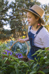 Fröhliches Kind mit Hut geht mit Weidenkorb im Garten spazieren und genießt den Sommerurlaub - ADSF17516