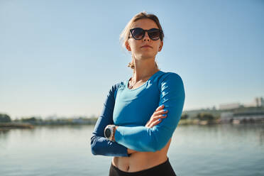 Selbstbewusste junge, fitte Frau in Sportkleidung und Sonnenbrille mit Fitness-Tracker, die am Flussufer steht und wegschaut - ADSF17520