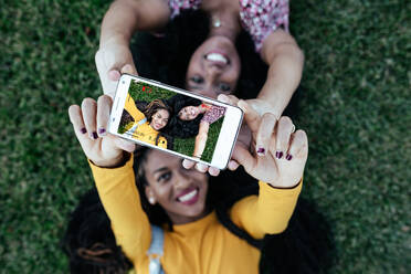 Draufsicht auf fröhliche beste Freundinnen, die zusammen auf grünem Gras liegen und ein Foto mit der Smartphone-Kamera machen, während sie das Wochenende genießen - ADSF17526