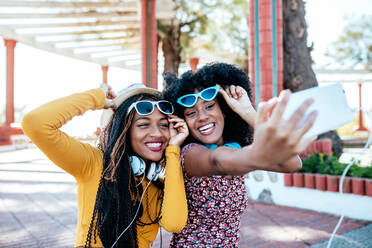 Smiling black woman with braids hugging cheerful African American female friend with curly hair and taking selfie together while standing on promenade in summer - ADSF17530
