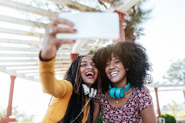 Smiling black woman with braids hugging cheerful African American female friend with curly hair and taking selfie together while standing on promenade in summer - ADSF17532