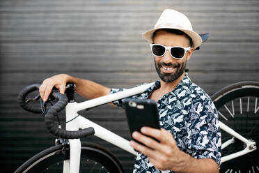 Cheerful stylish male standing on street with modern bicycle and taking photo on selfie camera of cellphone - ADSF17535