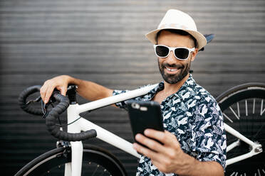 Fröhliche stilvolle männliche stehen auf der Straße mit modernen Fahrrad und nehmen Foto auf selfie Kamera des Handys - ADSF17535