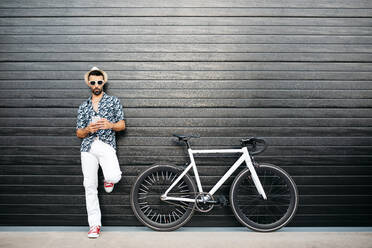 Seriöser Mann in modischer Kleidung, der in der Nähe eines Fahrrads an einer schwarzen Wand steht und auf einem Handy in der Stadt surft - ADSF17536