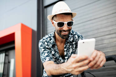 Delighted male in stylish outfit leaning on handlebar of bicycle and reading messages on social media via mobile phone in city - ADSF17537