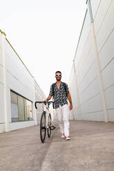 Niedriger Winkel von erfreut männlich in modischer Kleidung zu Fuß mit modernen Fahrrad entlang schmalen Straße und Blick nach vorn - ADSF17538
