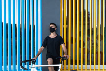 Ernster Mann mit Schutzmaske steht mit Fahrrad in der Nähe eines bunten Gebäudes in der Stadt und schaut weg während einer Coronavirus-Epidemie - ADSF17539