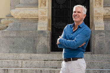 Delighted male in stylish clothes standing with crossed arms on stone stairway in city and looking away - ADSF17556