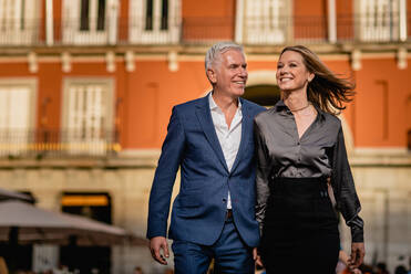 Elegant well dressed middle aged couple in stylish clothes smiling happily while walking together against blurred building on city street looking away - ADSF17560