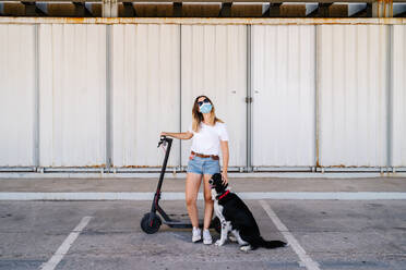 Teenagerin mit Sonnenbrille und Schutzmaske steht in der Nähe eines Elektrorollers und streichelt einen Hund auf einem Parkplatz - ADSF17582