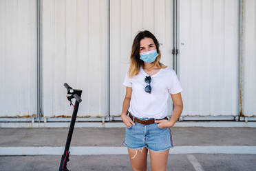 Ernste junge Studentin in Freizeitkleidung und medizinischer Maske schaut in die Kamera, während sie in der Nähe eines Elektrorollers an einer Metallwand steht - ADSF17583