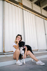 Full body side view of young female in casual outfit sitting with big dog near metal wall of urban building - ADSF17584