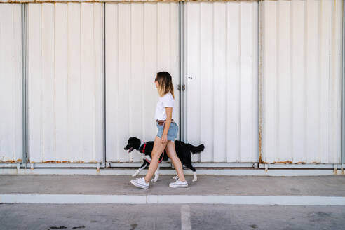Ganzkörper-Seitenansicht einer jungen Frau in Freizeitkleidung, die mit einem großen Hund in der Nähe einer Metallwand eines städtischen Gebäudes spazieren geht - ADSF17587