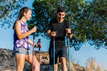 Niedriger Winkel eines glücklichen jungen Mannes und einer Frau in Sportkleidung mit Trekkingstöcken, die eine Pause einlegen, während sie auf einem Berghang stehen, während sie gemeinsam wandern und laufen, an einem Sommertag in der Natur - ADSF17596